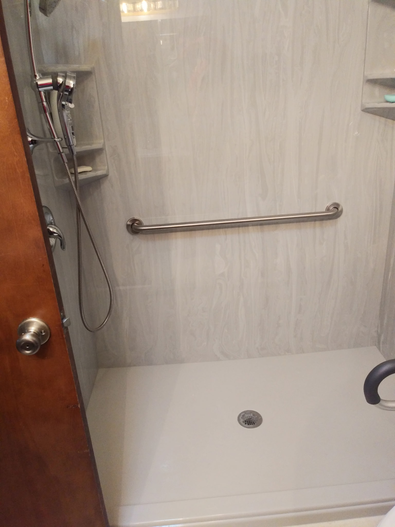 Shower with a horizontal grab bar, handheld shower on the left, wall niches, and a white shower base