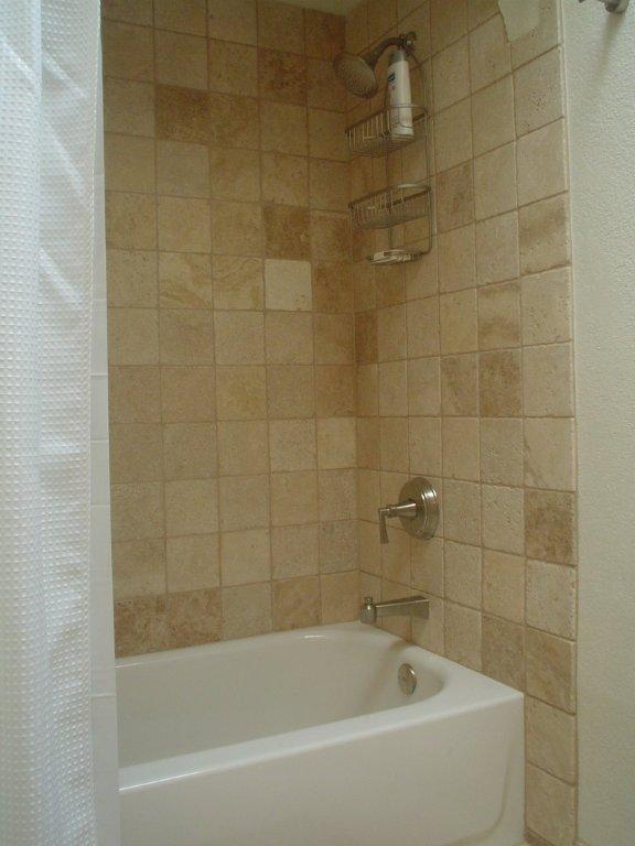 White tub with a travertine tile surround and wall-mounted shower